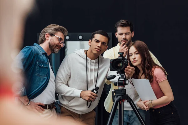 Enfoque selectivo de feliz director de arte cerca de compañeros de trabajo y cámara digital en el estudio de fotos - foto de stock