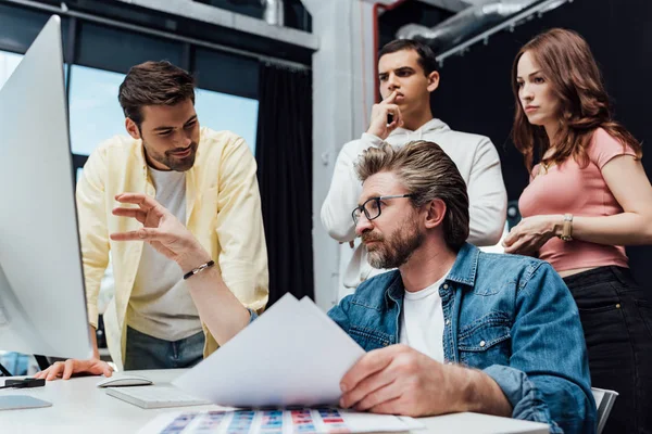 Heureux assistant souriant près directeur créatif gestuelle tout en regardant moniteur d'ordinateur — Photo de stock