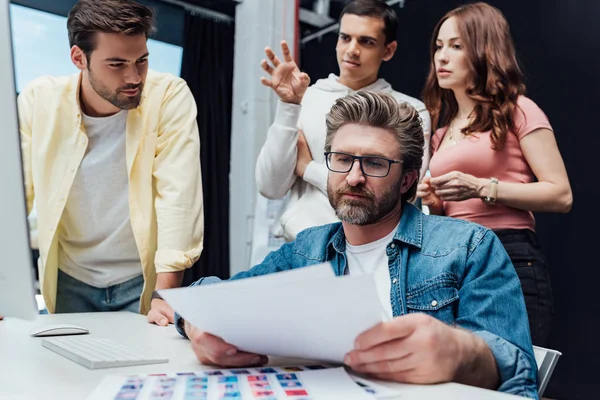 Selektiver Fokus eines gut aussehenden Art Directors, der Papiere in der Nähe von Assistenten betrachtet — Stockfoto