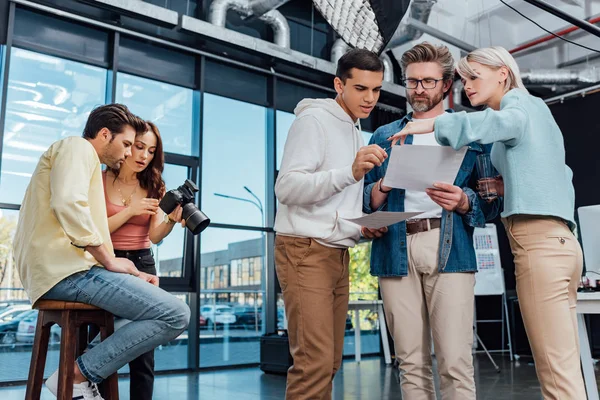 Director de arte en gafas mirando la foto cerca de compañeros de trabajo - foto de stock