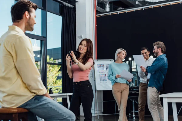 Selective focus of cheerful photographer taking photo of man near coworkers — Stock Photo