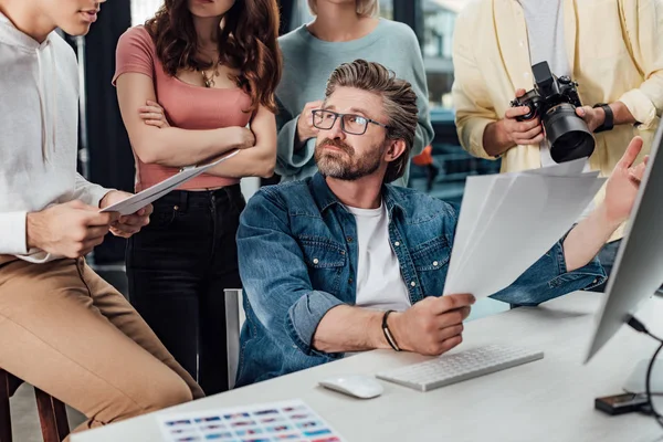 Beau directeur artistique tenant des photos et regardant des assistants — Photo de stock