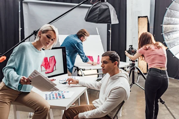 Attractive art director looking at photos near computer monitor and assistant — Stock Photo