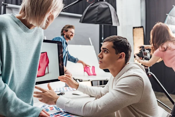 Selective focus of handsome man gesturing near woman and computer monitor in photo studio — Stock Photo