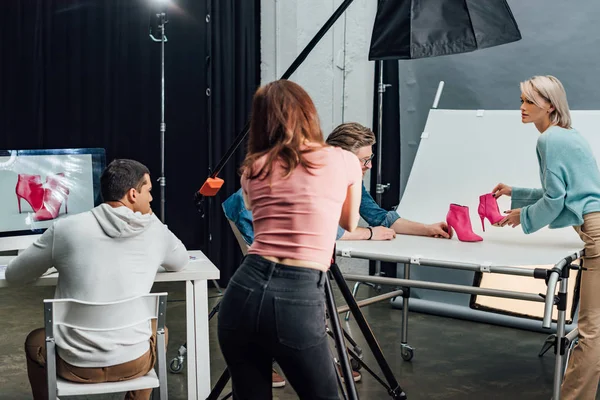 Visão traseira do fotógrafo tirando foto de sapatos rosa perto de colegas de trabalho no estúdio de fotografia — Fotografia de Stock