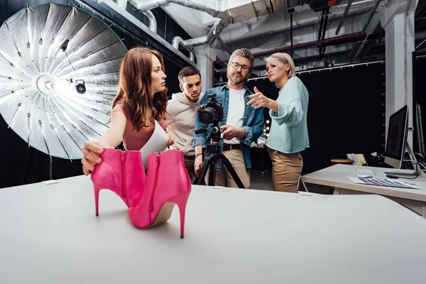 Mujer tocando zapatos rosados cerca de compañeros de trabajo durante sesión de fotos - foto de stock