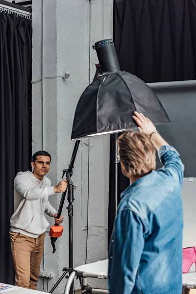 Apuesto asistente celebración reflector cerca de director de arte en estudio de fotos - foto de stock