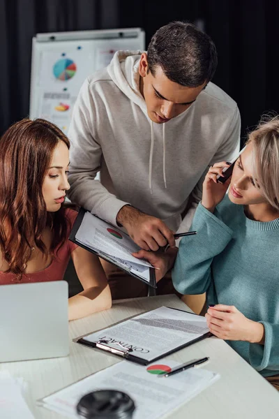 Enfoque selectivo de asistente guapo sujetando portapapeles cerca de director creativo y compañero de trabajo - foto de stock