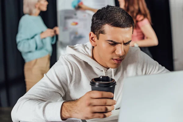 Enfoque selectivo de guapo director de arte sosteniendo taza de papel mientras se utiliza el ordenador portátil - foto de stock