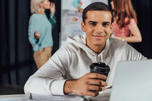 Selective focus of happy art director holding paper cup while using laptop — Stock Photo