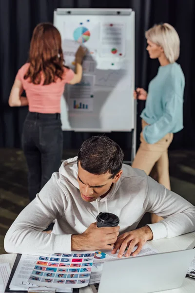 Selektiver Fokus eines gut aussehenden Art Directors, der Pappbecher hält, während er Collage in Laptopnähe betrachtet — Stockfoto