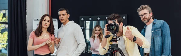 Fotografía panorámica del fotógrafo tomando fotos cerca del director de arte en gafas y mostrando el pulgar hacia arriba - foto de stock