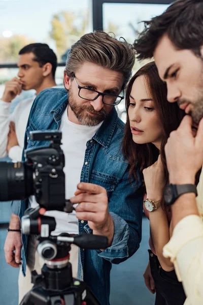 Enfoque selectivo del director de arte barbudo apuntando con el dedo a la cámara digital cerca de los asistentes - foto de stock