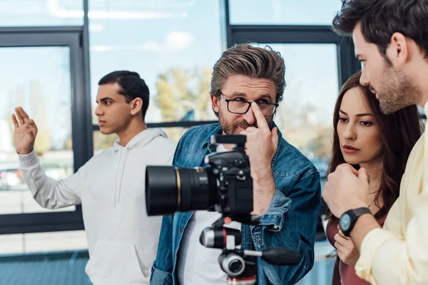 Foco seletivo do diretor de arte tocando óculos e olhando para assistente perto da câmera digital — Fotografia de Stock