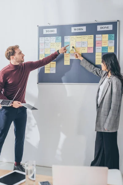 Selective focus of handsome scrum master pointing with finger at sticky note with db design letters near asian coworker — Stock Photo
