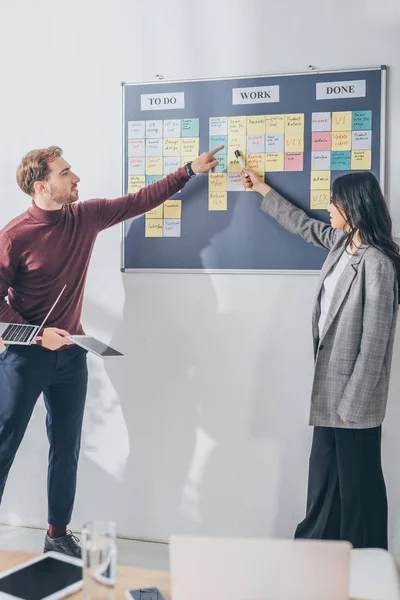 Selektiver Fokus eines gutaussehenden Gedrängemeisters, der mit dem Finger auf eine klebrige Note mit db-Design-Buchstaben in der Nähe asiatischer Mitarbeiter zeigt — Stockfoto