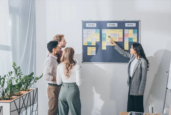 Attractive asian scrum master pointing at db design letters on sticky note near businessmen and businesswoman — Stock Photo