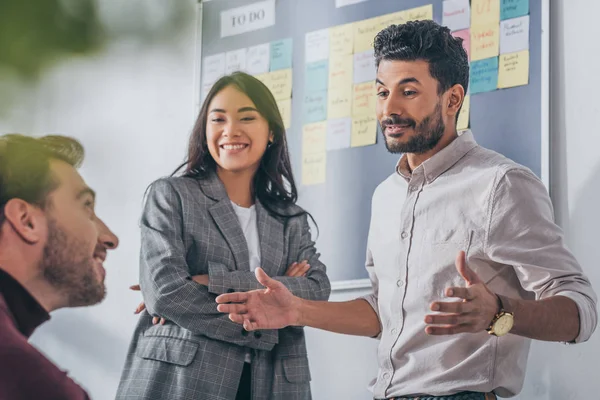 Enfoque selectivo de compañeros de trabajo multiculturales alegres hablando en la oficina - foto de stock