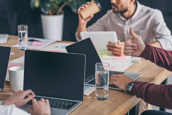 Cropped view of man showing thumb up near laptops and coworkers — Stock Photo