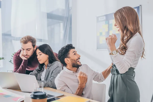 Excited mixed race businessman and businessman celebrating near coworkers in office — Stock Photo