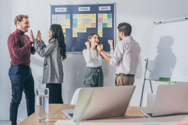 Excited mixed race businessman and happy businessman celebrating near coworkers giving high five in office — Stock Photo