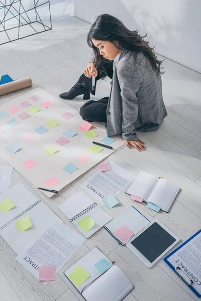 Hermosa asiático scrum maestro sentado en piso cerca de documentos con letras - foto de stock