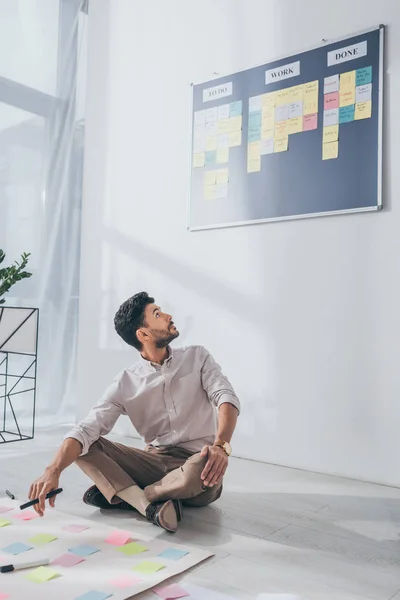 Mixed race scrum master sitting on floor near sticky notes and looking on board — Stock Photo