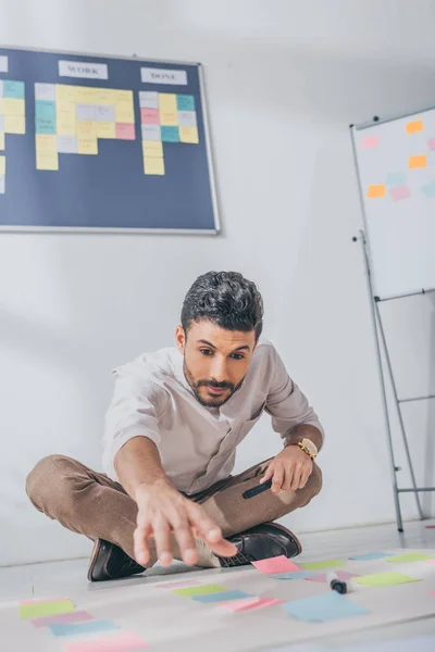 Selektiver Fokus des Mixed Race Scrum Master, der mit ausgestreckter Hand in der Nähe klebriger Noten auf dem Boden sitzt — Stockfoto