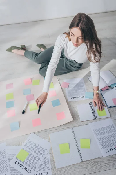 Blick von oben auf attraktive Geschäftsfrau, die auf dem Boden sitzt und Filzstift in der Nähe klebriger Notizen hält — Stockfoto