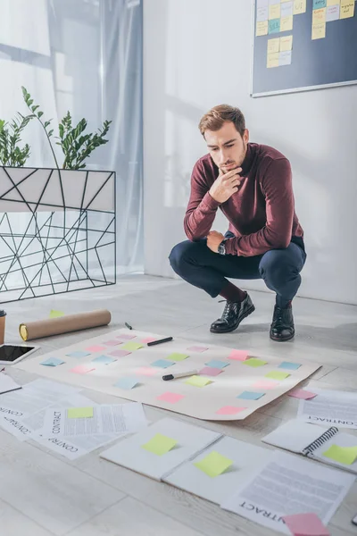 Nachdenklicher Gedrängemeister, der sitzt und das Gesicht berührt, während er auf Vertrag und klebrige Notizen auf dem Boden blickt — Stockfoto