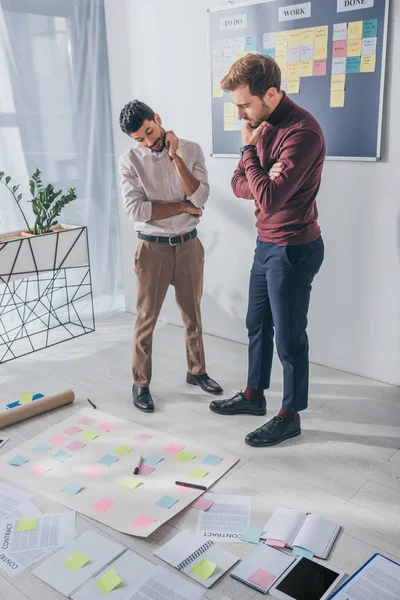 Maestros de scrum multiculturales pensativos de pie y mirando notas adhesivas en el suelo - foto de stock