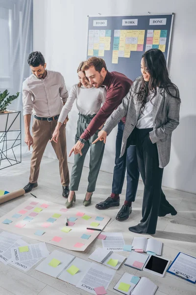 Happy multicultural scrum masters pointing with fingers at sticky notes on floor — Stock Photo