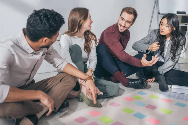 Happy multiccultural scrum masters sitting on floor near sticky notes — стоковое фото