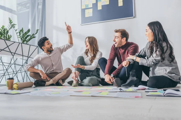 Happy multicultural scrum masters sitting on floor and looking at mixed race coworker with idea — Stock Photo