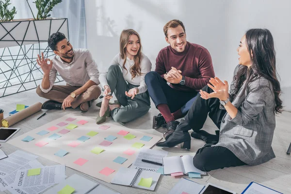 Glücklich gemischte Rasse Geschäftsmann zeigt ok Zeichen in der Nähe Mitarbeiter auf dem Boden sitzen — Stockfoto