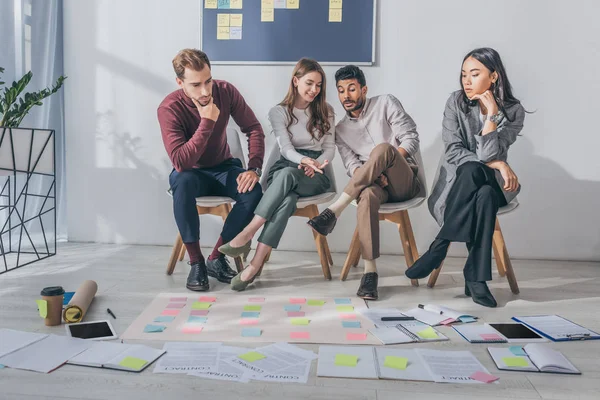 Atractiva mujer de negocios señalando con la mano notas adhesivas en el suelo cerca de compañeros de trabajo multiculturales - foto de stock