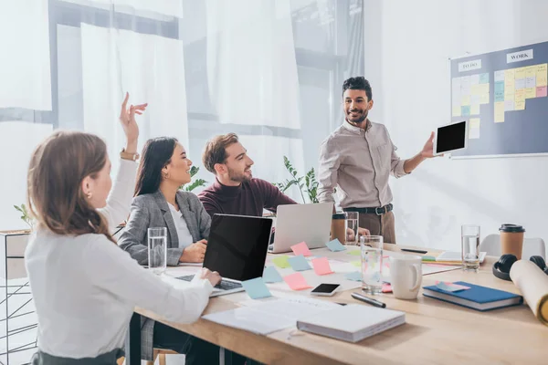 Bi-racial scrum master looking at businesswoman with raising hand near coworkers — Stock Photo