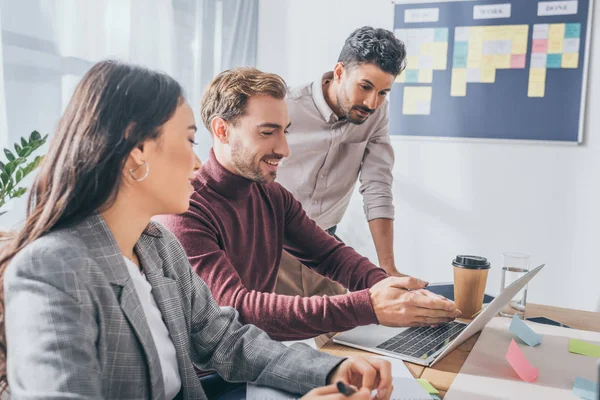 Selektiver Fokus des fröhlichen Geschäftsmannes, der mit der Hand auf Laptop neben asiatischer Geschäftsfrau und Mischlingskollegin zeigt — Stockfoto