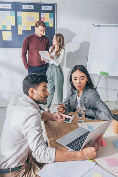 Selective focus of mixed race businessman pointing with finger at laptop near asian coworker — Stock Photo