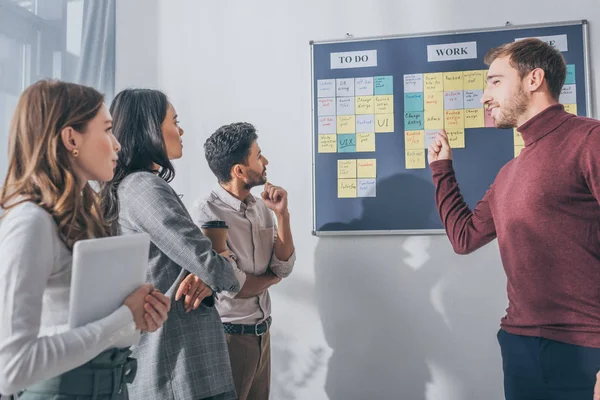 Selective focus of handsome scrum master pointing with finger at board near multicultural coworkers — Stock Photo