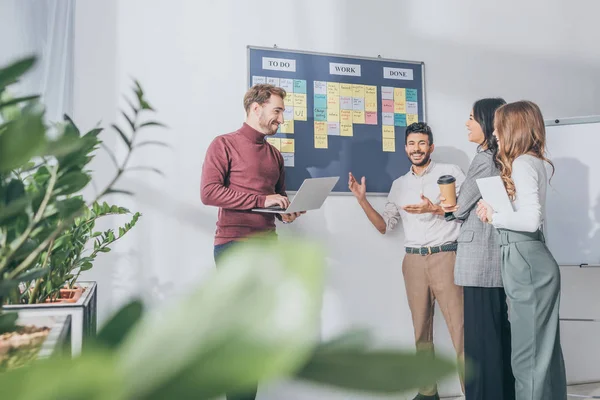 Selective focus of scrum master using laptop near board with letters and multicultural coworkers — Stock Photo