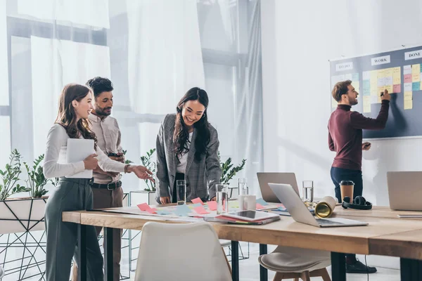 Alegres empresarias multiculturales cerca de hombre de negocios y gadgets en la oficina - foto de stock