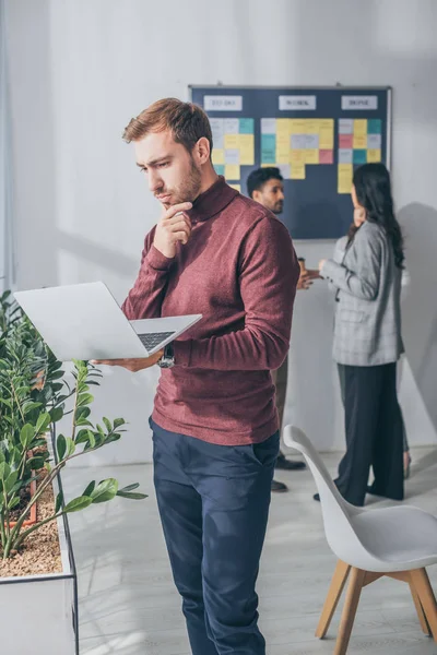 Enfoque selectivo de maestro scrum guapo utilizando portátil cerca de compañeros de trabajo multiculturales - foto de stock