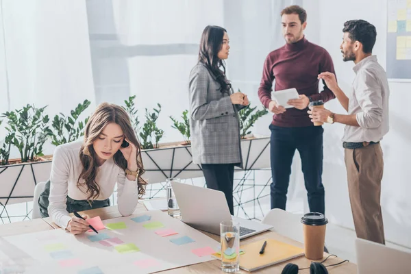 Selective focus of attractive businesswoman writing on sticky note near multicultural coworkers — Stock Photo