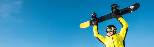 Tiro panorâmico de desportista feliz no capacete segurando snowboard acima da cabeça — Fotografia de Stock