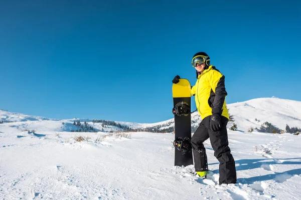 Desportista feliz no capacete segurando snowboard e de pé na neve branca nas montanhas — Fotografia de Stock