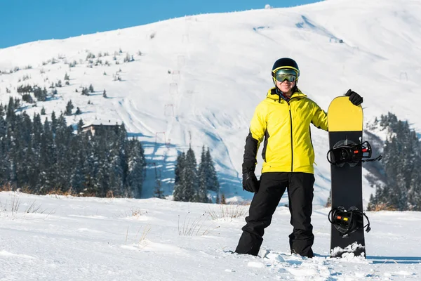 Desportista alegre no capacete segurando snowboard e de pé na neve branca nas montanhas — Fotografia de Stock