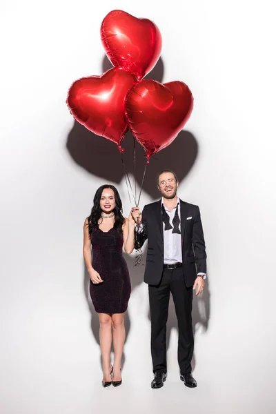 Hermosa pareja sonriente sosteniendo globos en forma de corazón rojo en día de San Valentín en blanco - foto de stock