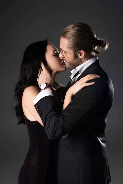 Sensual pareja en traje negro y vestido besos aislados en gris - foto de stock