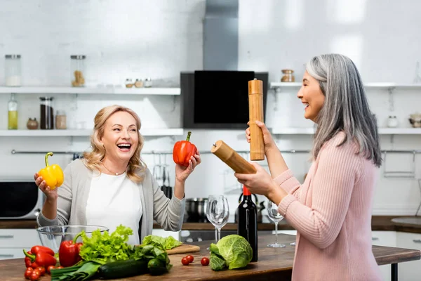 Sonriente mujer sosteniendo pimientos y su asiático amigo sosteniendo pimienta molino y sal molino - foto de stock